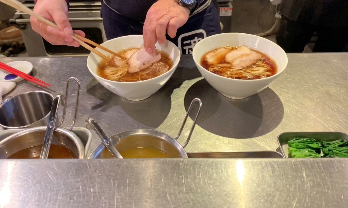 Ein Koch legt Scheiben von gegartem Schweine-Chashu mit Stäbchen auf die arrangierten Ramen-Nudeln.