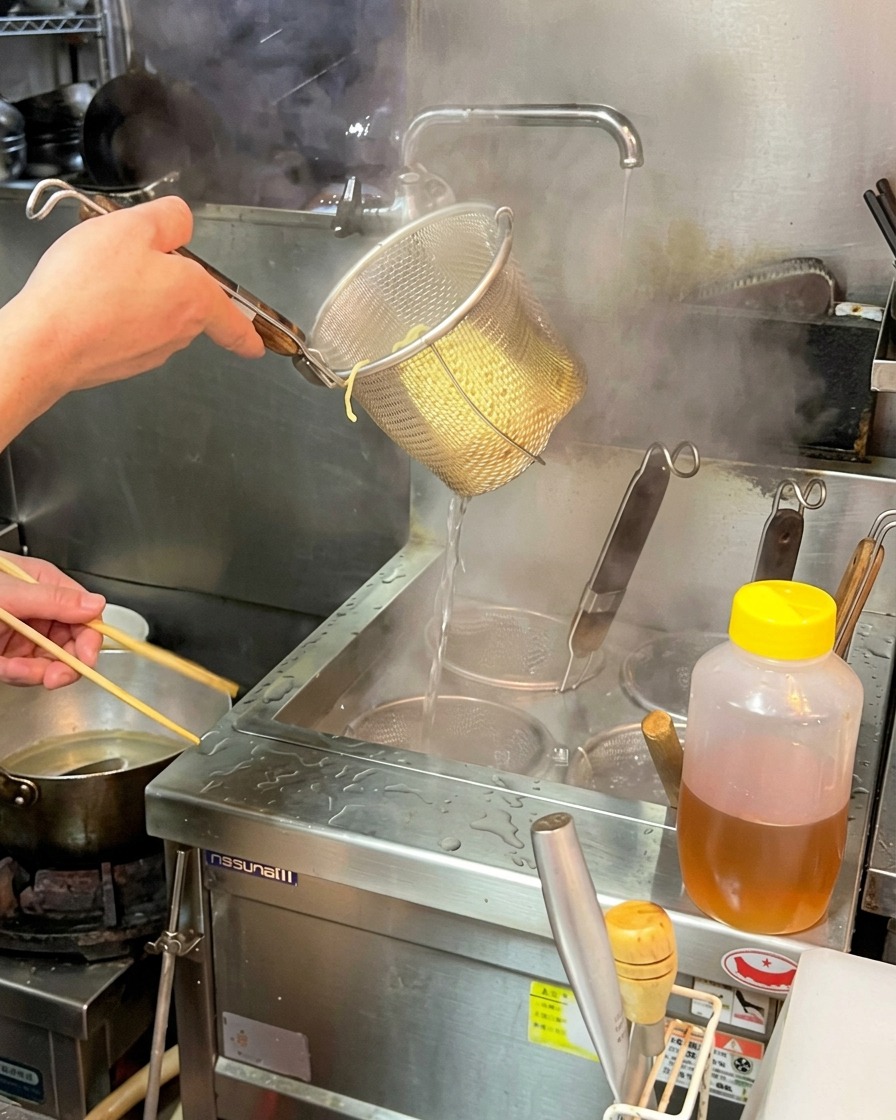 Ein Koch schüttelt kräftig ein Sieb mit gegarten Ramen-Nudeln ab.