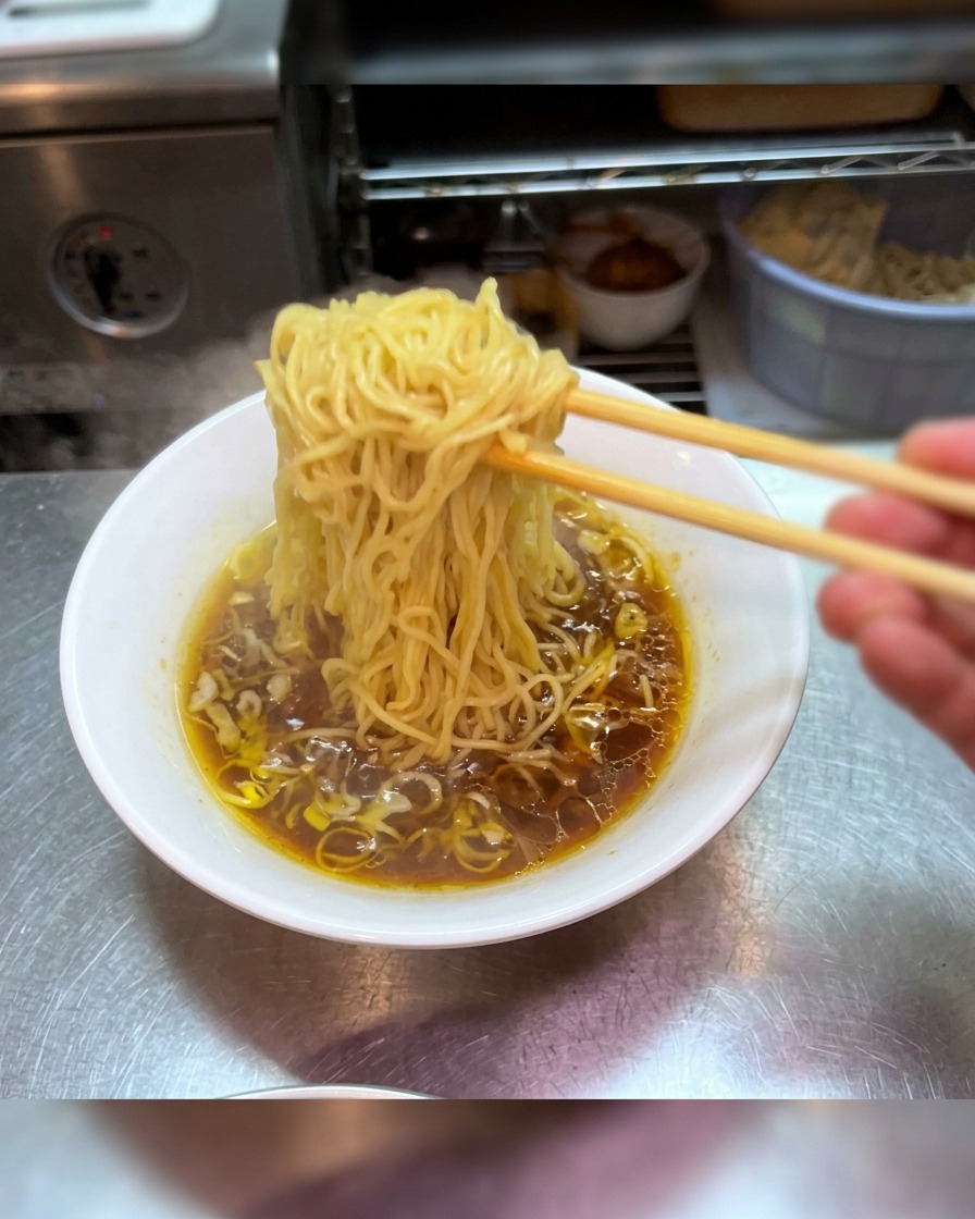 Stäbchen heben ein Bündel frischer Ramen-Nudeln hoch über eine Schüssel mit dunkler Shoyu-Brühe.