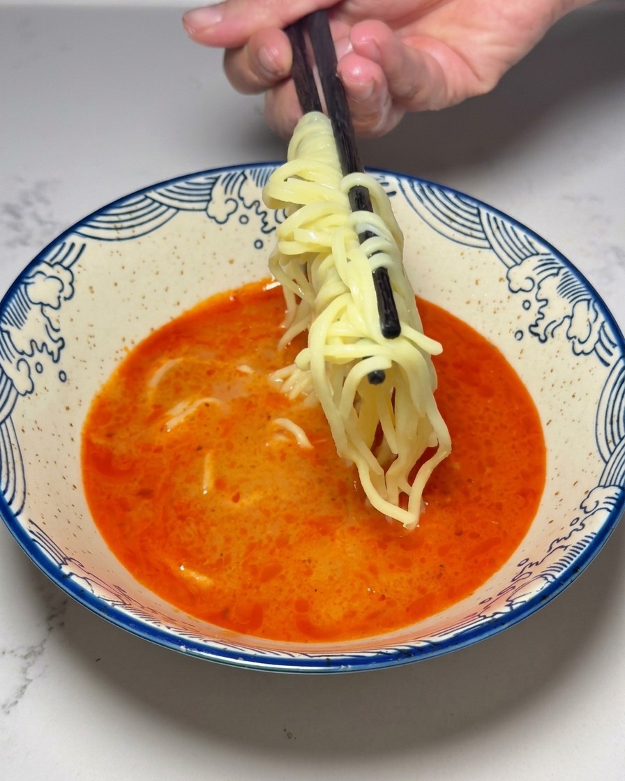 Gelbe Ramen-Nudeln werden mit Stäbchen in die rote Suppe gelegt.