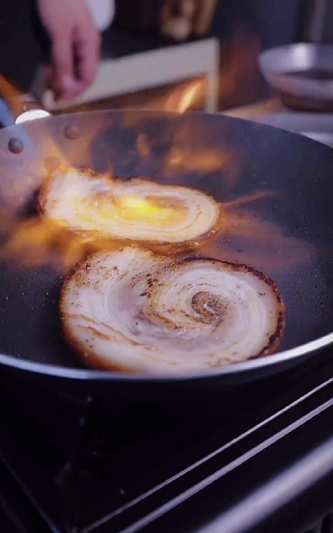 Zwei Chashu-Scheiben in einer Pfanne werden mit der hellorangen Flamme eines Küchenbrenners bearbeitet.