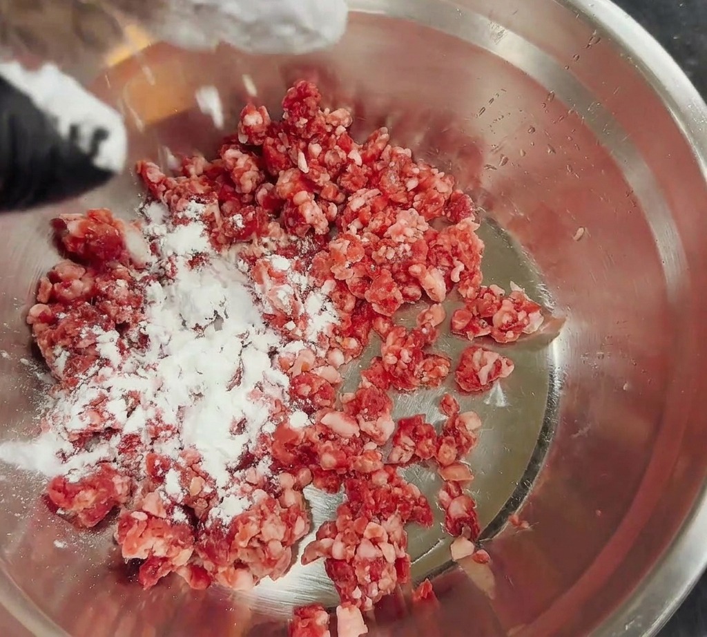 Maisstärke wird über frisches Rinderhackfleisch in einer Edelstahlschüssel gestreut.