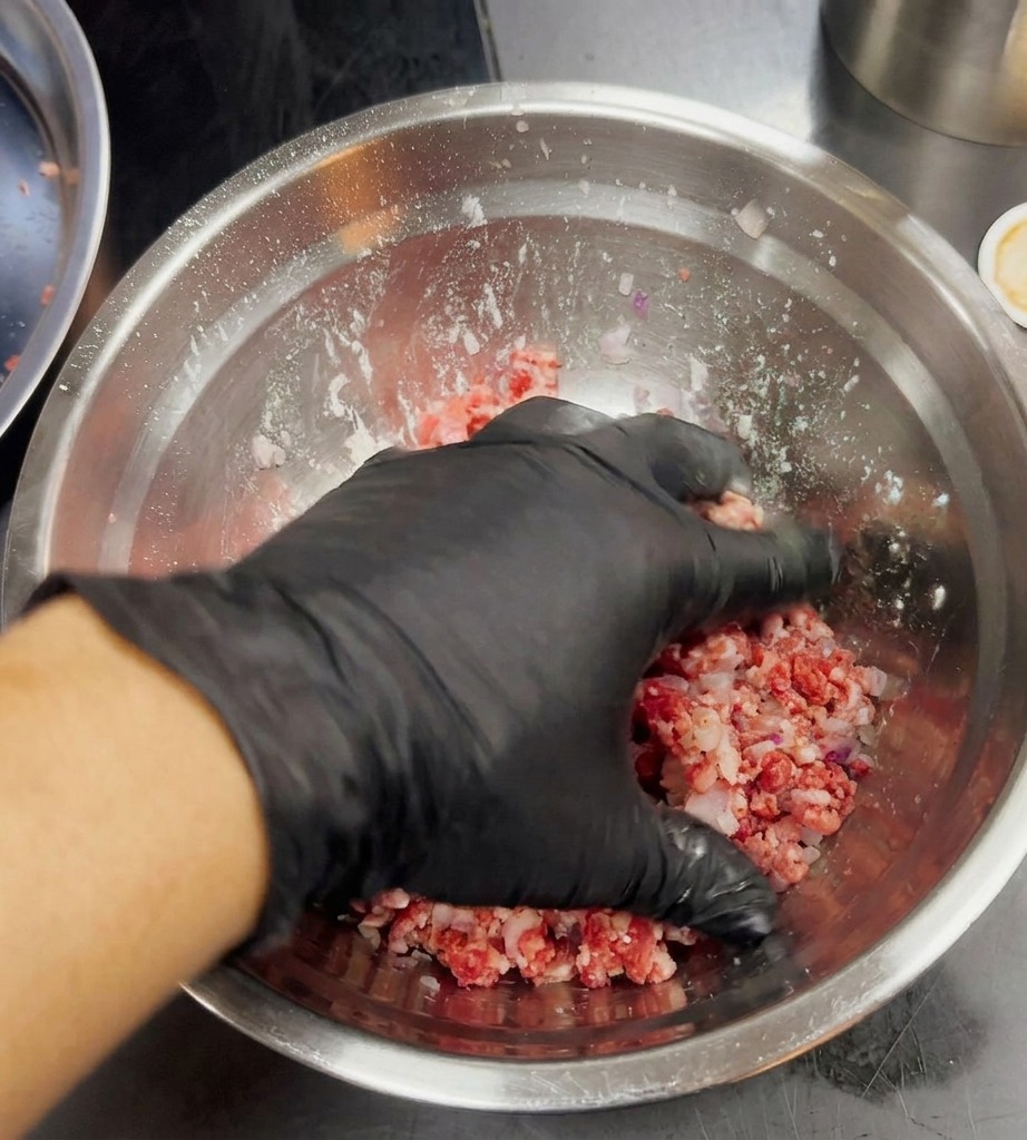 Eine Hand in einem schwarzen Handschuh mischt vorsichtig Hackfleisch und gemahlene Zwiebeln.