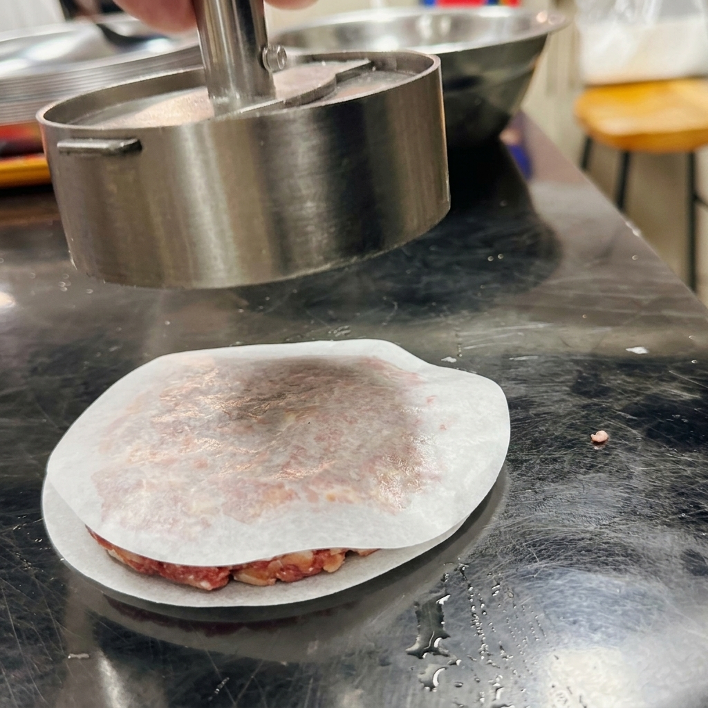 Eine Burgerpresse aus Metall formt das Hackfleisch auf Backpapier zu einem runden Patty.