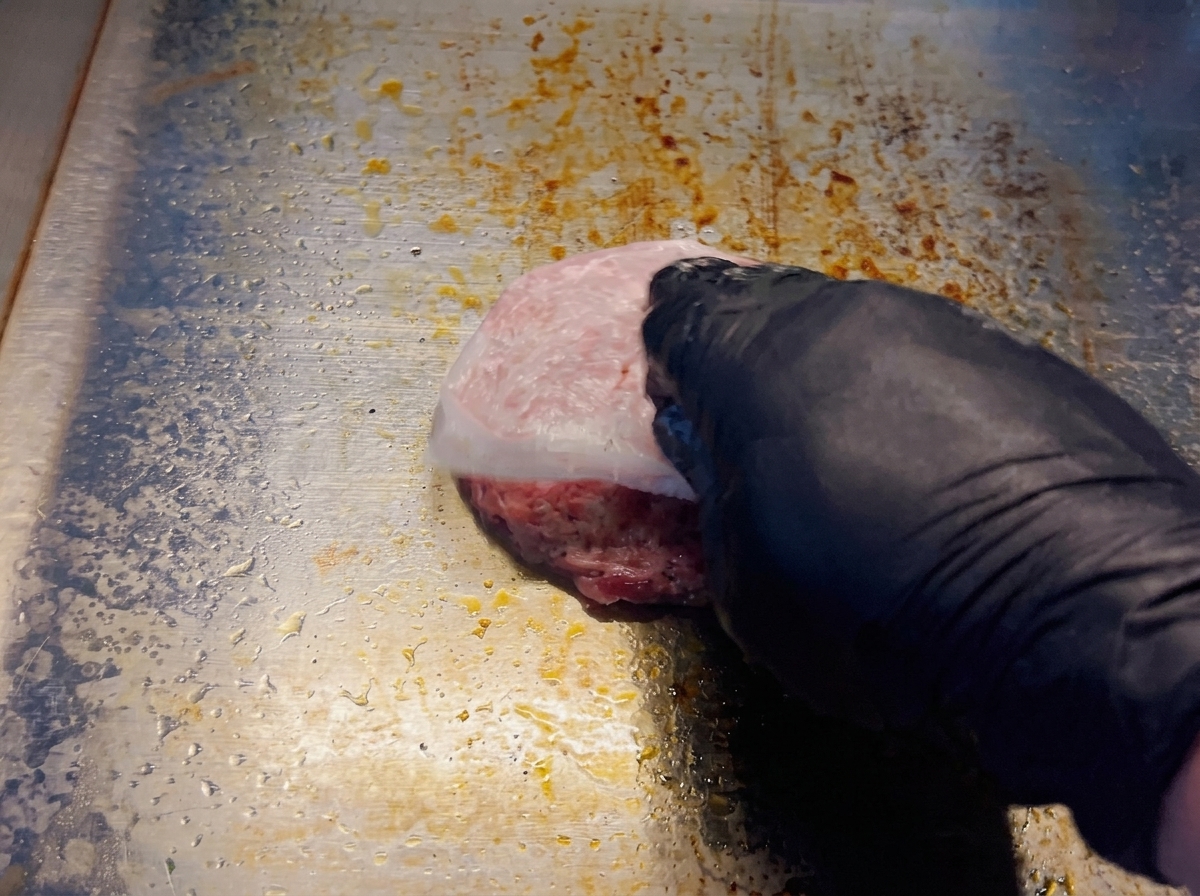 Eine Hand zieht das Backpapier von einem rohen Patty auf einer heißen Grillplatte ab.