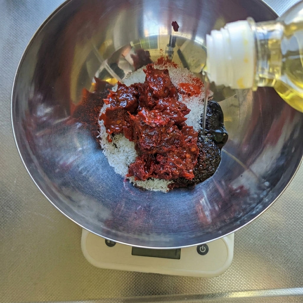 Salatöl wird in eine Edelstahlschüssel gegossen, in der sich bereits dunkelrote Chilipaste, weißer Zucker und Gewürze befinden, alles steht auf einer Waage.