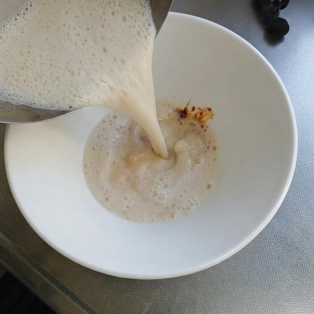 Heiße, cremig-weiße Tonkotsu-Brühe wird aus einem Metalltopf in eine weiße Schüssel gegossen, in der sich eine kleine Menge dunkelroter Chilipaste befindet.