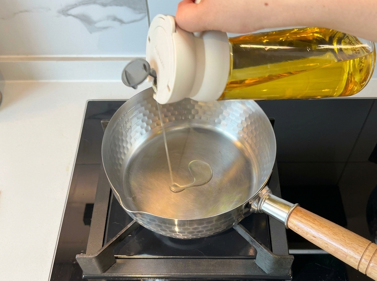 Eine Hand gießt Speiseöl aus einer Flasche in einen sauberen Edelstahltopf auf dem Herd.