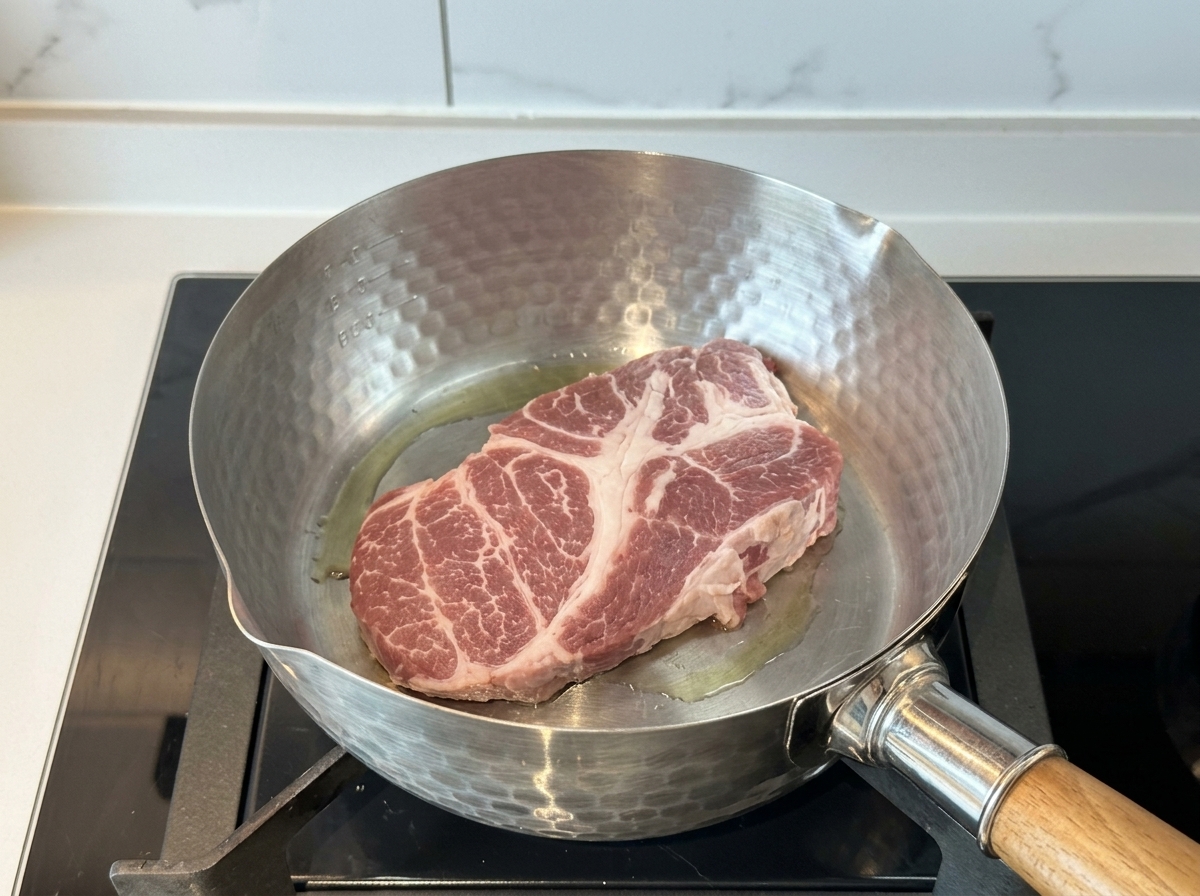 Ein dickes, rohes Stück Schweineschulter brät in einem heißen Edelstahltopf mit etwas Öl.