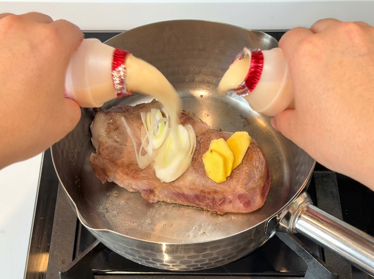 Hände gießen zwei kleine Fläschchen Joghurtgetränk über ein angebratenes Stück Schweinefleisch mit Zwiebeln und Ingwer in einem Edelstahltopf.