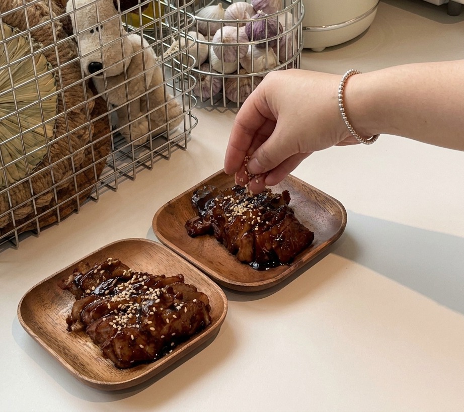 Eine Hand streut weißen Sesam über in Scheiben geschnittenes, glasiertes Schweinefleisch, das auf zwei kleinen, quadratischen Holztellern angerichtet ist.