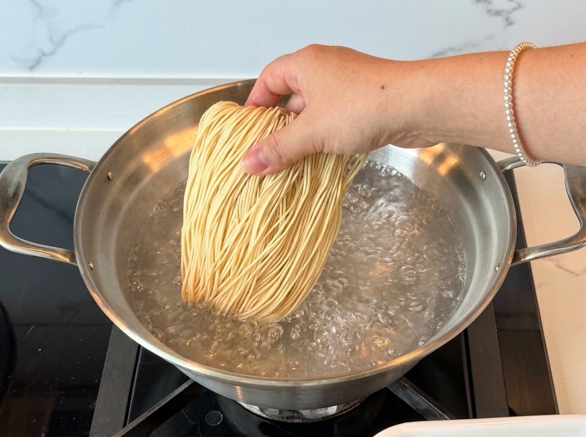 Eine Hand gibt ein Bündel gerader, getrockneter Ramen-Nudeln in einen Edelstahltopf mit sprudelnd kochendem Wasser.