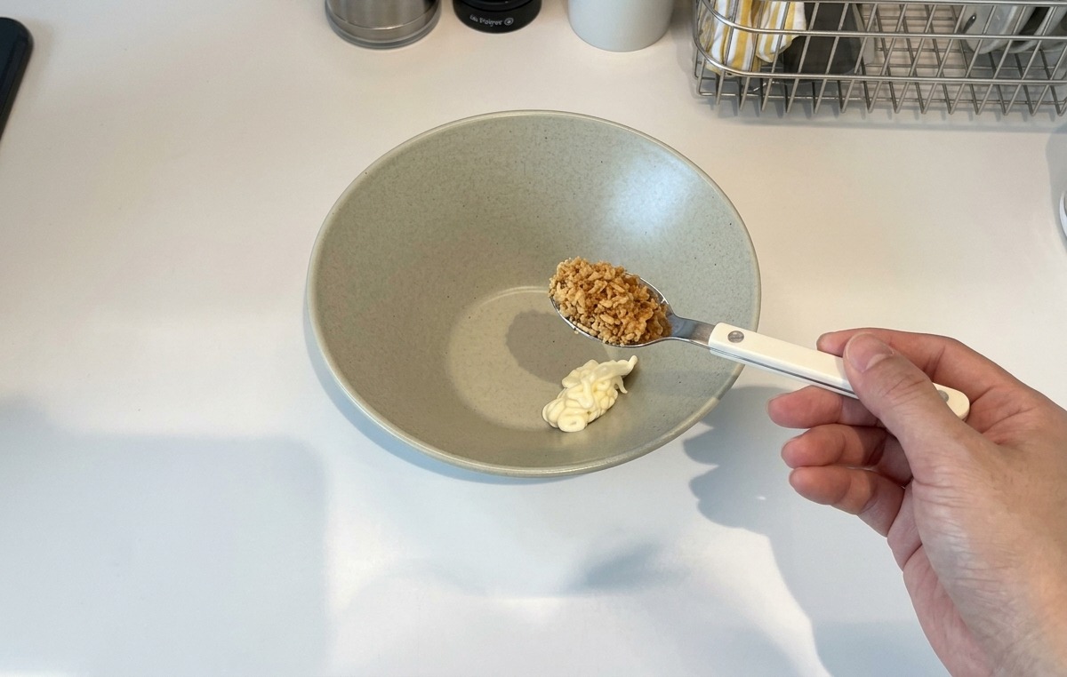 Eine Hand hält einen Löffel mit knusprig geröstetem Knoblauch über eine Keramikschale, in der sich ein Klecks Mayonnaise befindet.
