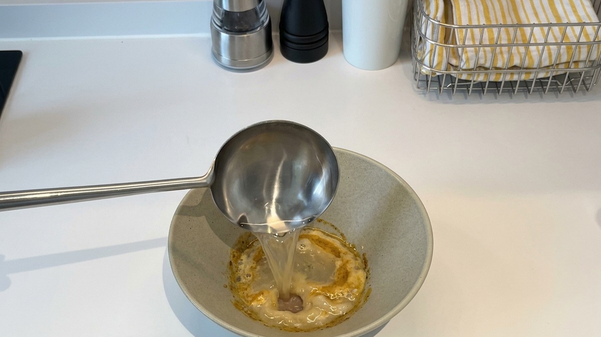Eine Schöpfkelle aus Edelstahl gießt heißes Wasser in eine Keramikschüssel mit einer dicken, gelben Würzpaste.