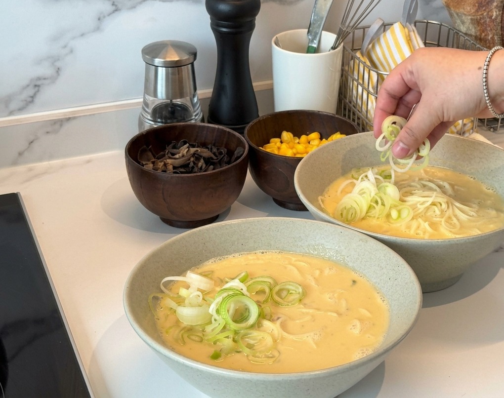 Eine Hand streut dünn geschnittene Frühlingszwiebeln über zwei Schüsseln mit cremiger, leicht gelblicher Ramen-Brühe.