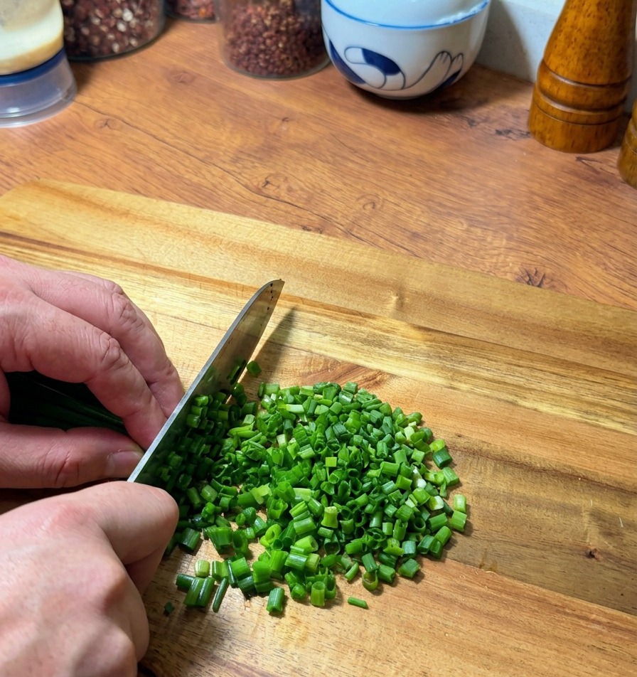 Hände schneiden frische grüne Frühlingszwiebeln auf einem Holzbrett mit einem Kochmesser.