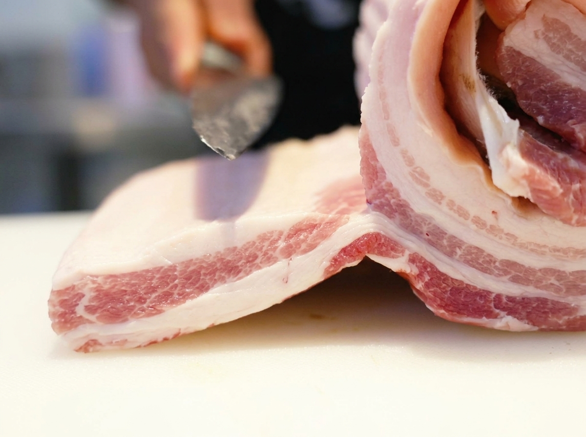 Ein scharfes Messer schneidet in ein dickes Stück rohen Schweinebauch auf einem weißen Schneidebrett.