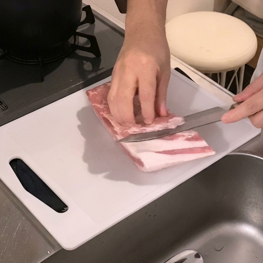 Hände schneiden die Innenseite eines rohen Schweinebauchs auf einem weißen Schneidebrett mit einem Messer ein.