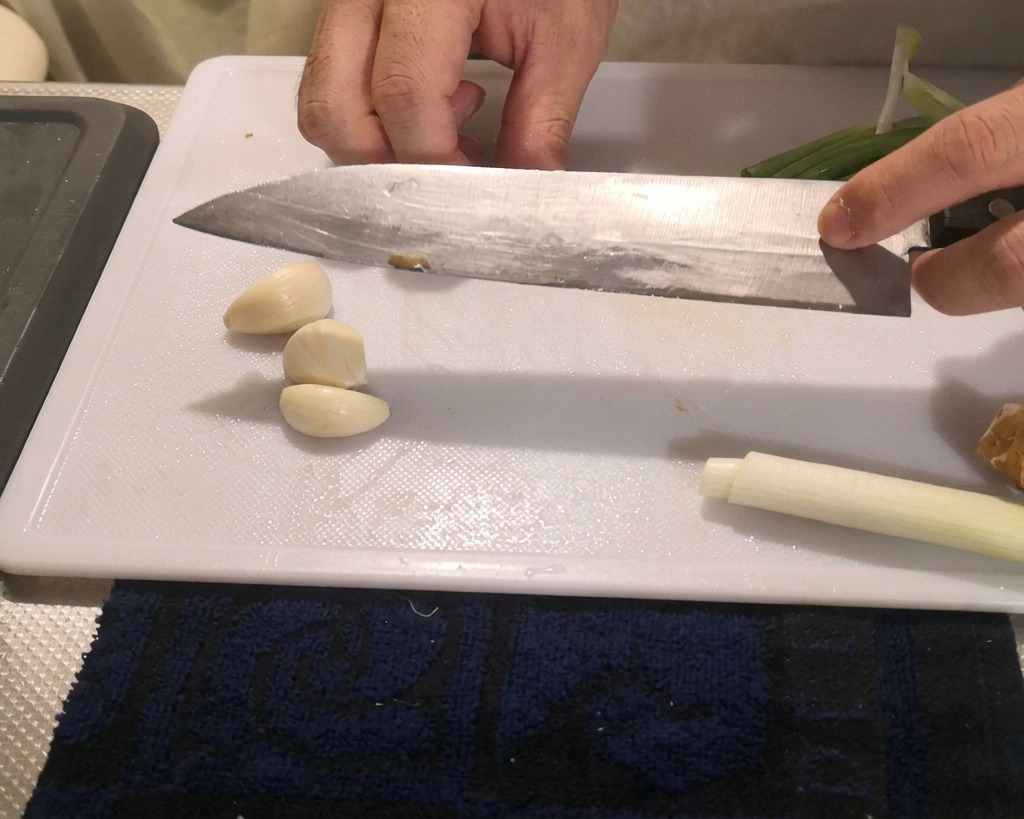 Eine Person drückt mit der flachen Seite eines großen Kochmessers auf geschälte Knoblauchzehen auf einem Brett.