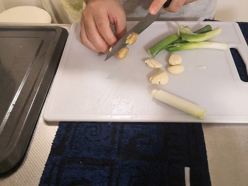 Ein Koch schneidet ein Stück frischen Ingwer in dicke Scheiben auf einem Brett neben Zwiebeln und Knoblauch.