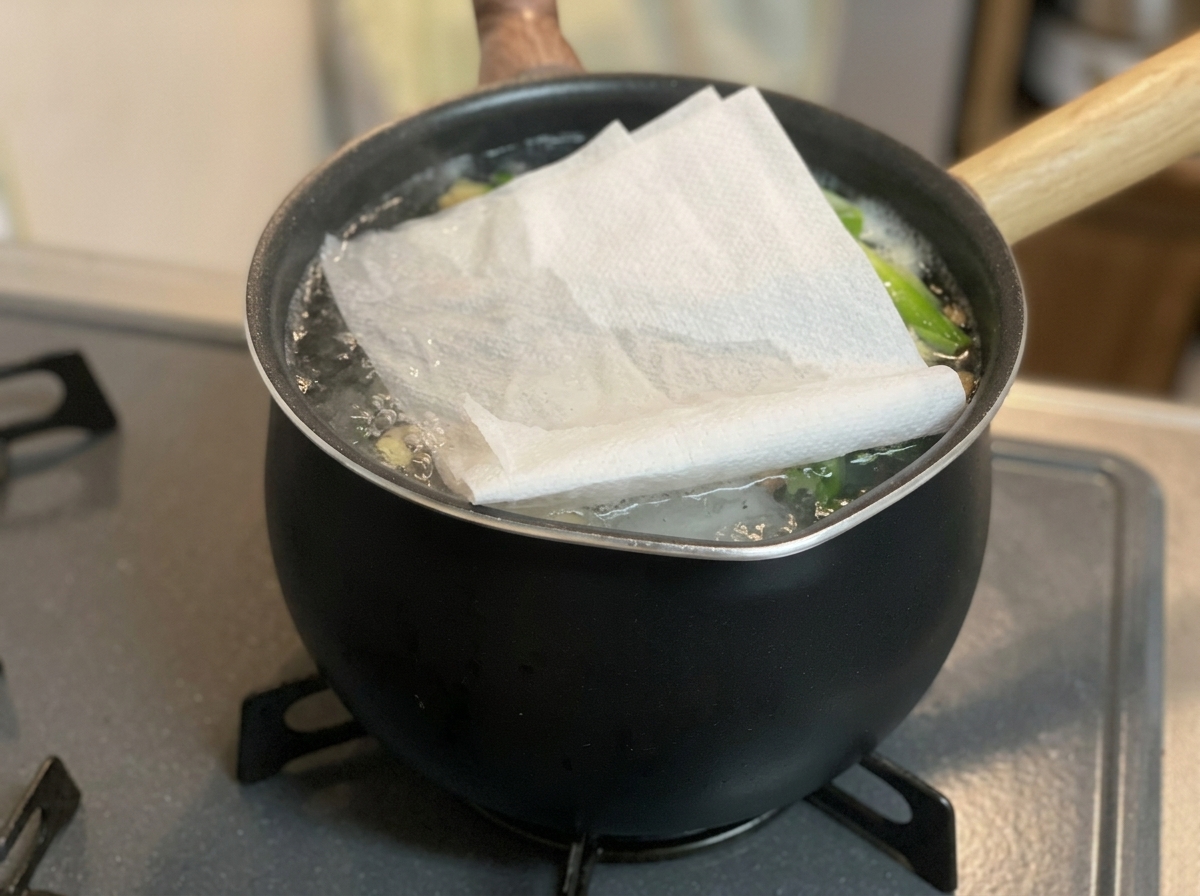 Ein gefaltetes weißes Papiertuch liegt direkt auf der Oberfläche eines köchelnden Suds in einem schwarzen Topf.