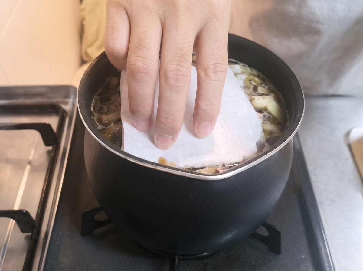 Eine Hand legt ein weißes Papiertuch auf einen köchelnden Topf mit dunkler Brühe.
