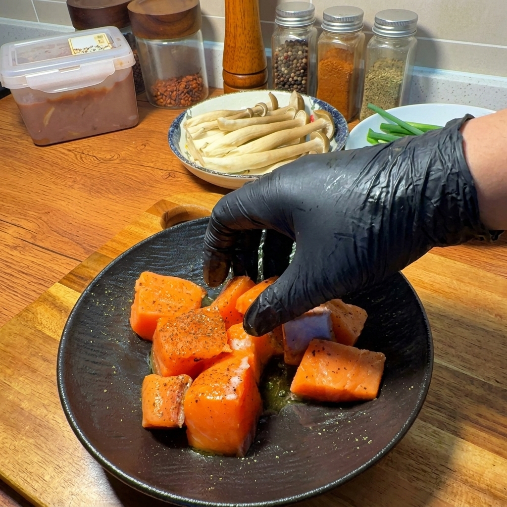 Eine Hand vermengt die Lachswürfel vorsichtig mit Gewürzen und Olivenöl auf einem dunklen Teller.