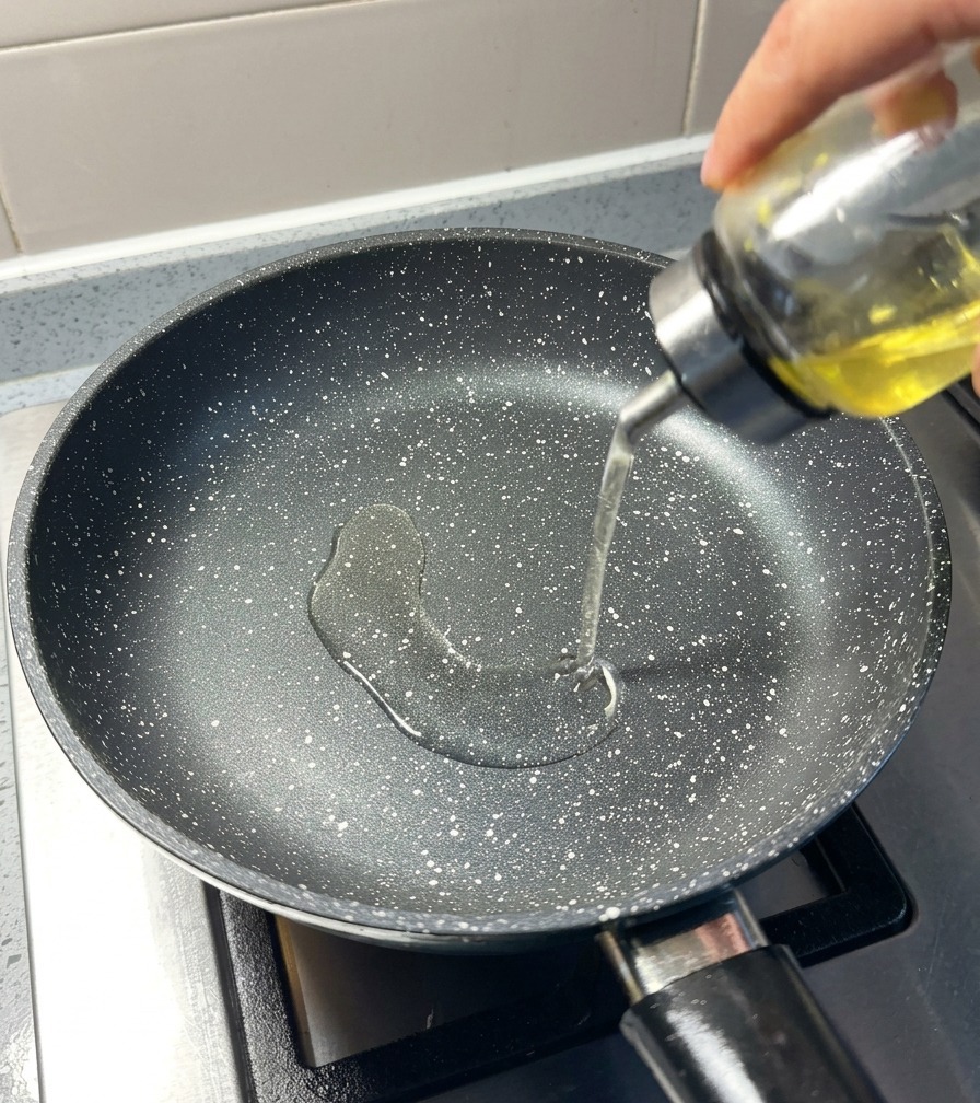 Helles Pflanzenöl wird aus einem Glasbehälter in eine erhitzte, schwarz gesprenkelte Pfanne gegossen.