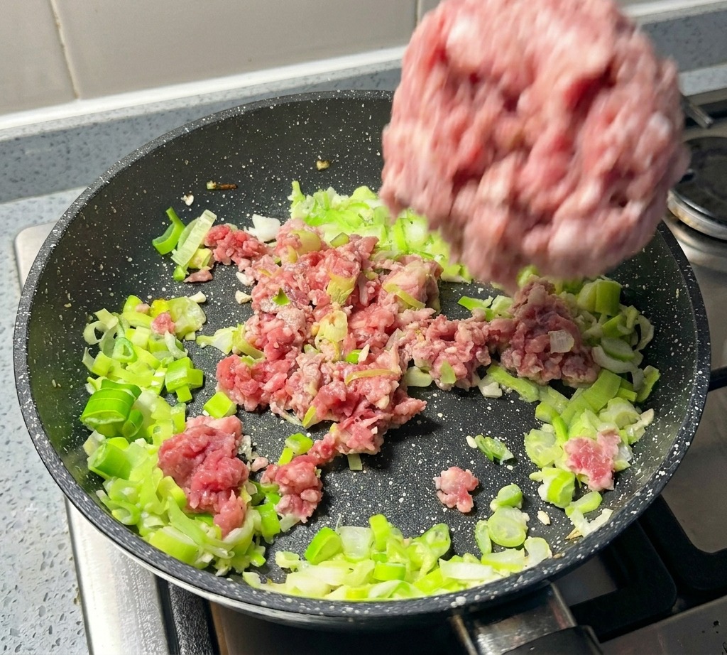 Eine Portion rohes Schweinehackfleisch wird zu den angebratenen Frühlingszwiebeln in die Pfanne gegeben.