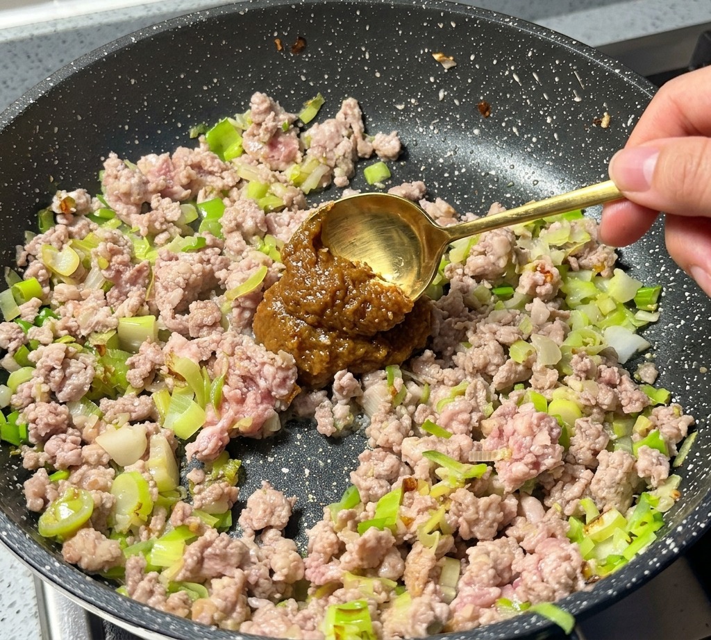Ein goldener Löffel gibt einen Klecks dunkle Misopaste zum gebräunten Hackfleisch in der Pfanne.