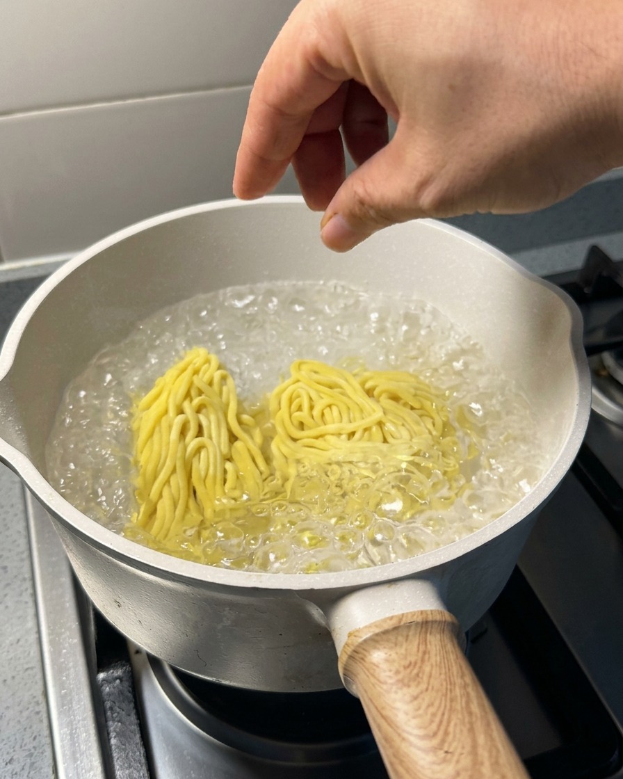 Zwei Portionen rohe gelbe Ramen-Nudeln werden in einen Topf mit sprudelnd kochendem Wasser gegeben.