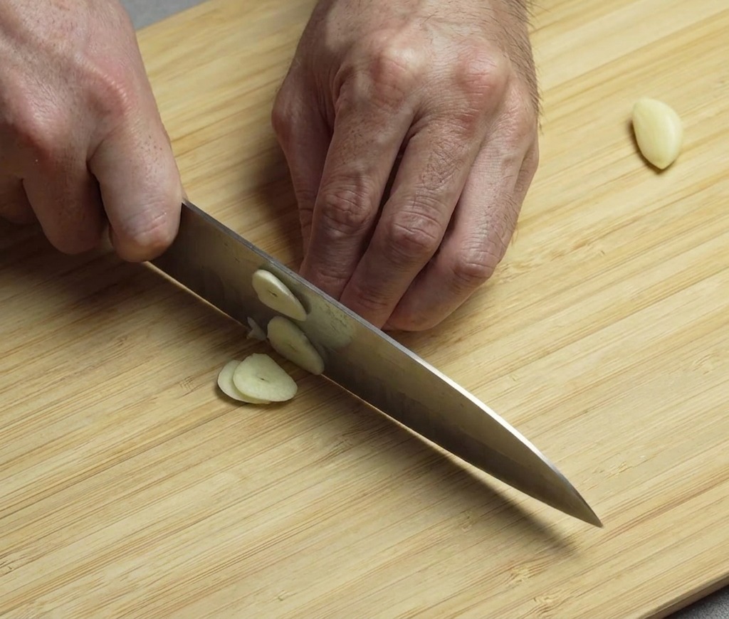 Hände nutzen ein Messer, um geschälte Knoblauchzehen auf einem Holzbrett fein zu hacken.