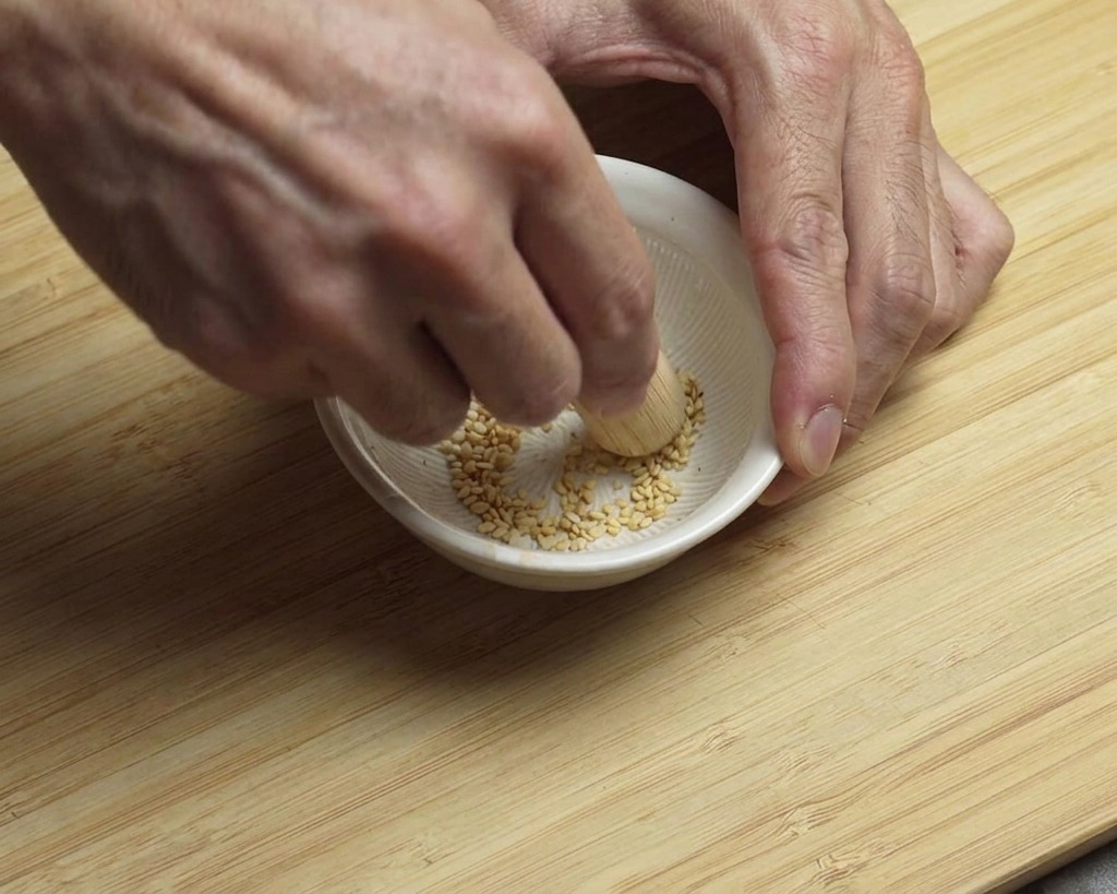 Zwei Hände zerstoßen weißen Sesam mit einem Holzstößel in einer weißen Reibschale.