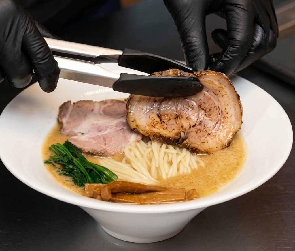 Eine Zange legt eine große Scheibe Chashu-Schweinebauch in die Ramen.