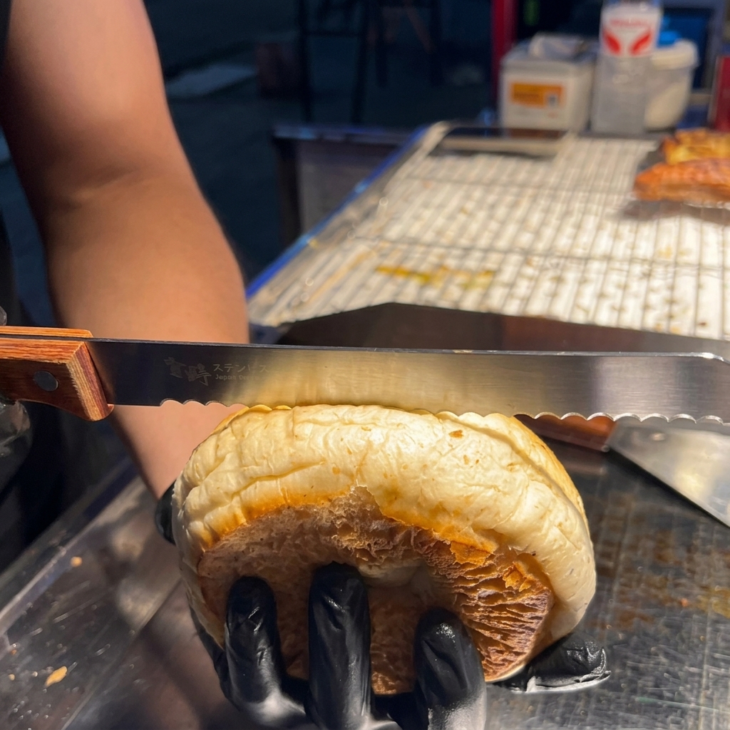 Eine Hand schneidet mit einem Brotmesser ein Hamburger-Brötchen mit Sesam vorsichtig waagerecht durch.