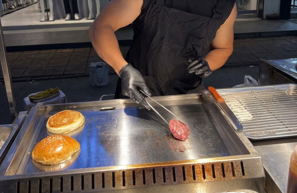 Fleischkugel aus Rinderhack wird mit einer Grillzange auf eine rauchend heiße Grillplatte gelegt.