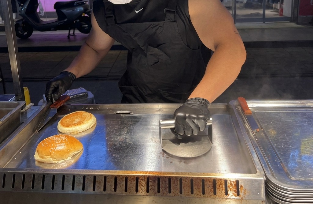 Eine schwere Metall-Burgerpresse drückt eine Kugel Hackfleisch auf einer heißen Platte extrem flach.