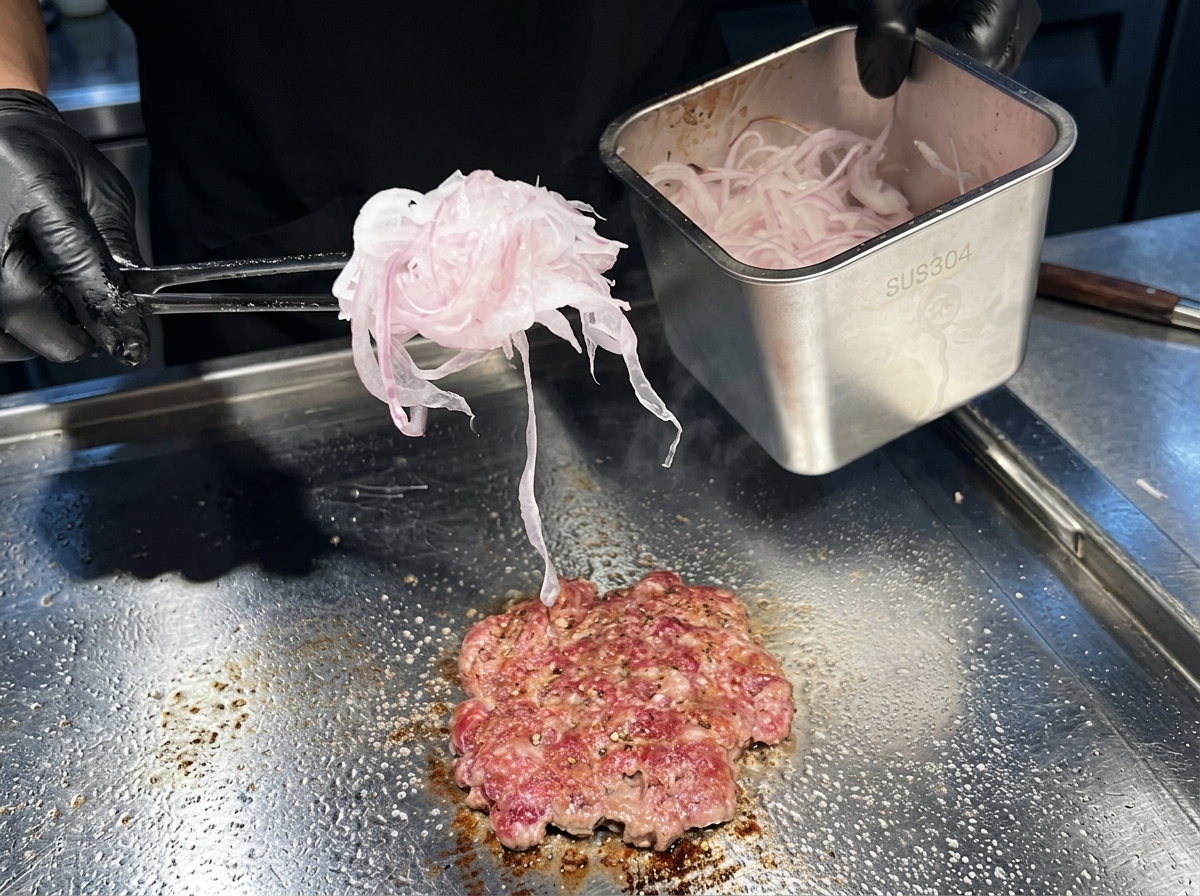 Fein geschnittene rote Zwiebeln werden auf das gärende Fleischpatty auf der Grillplatte gegeben.