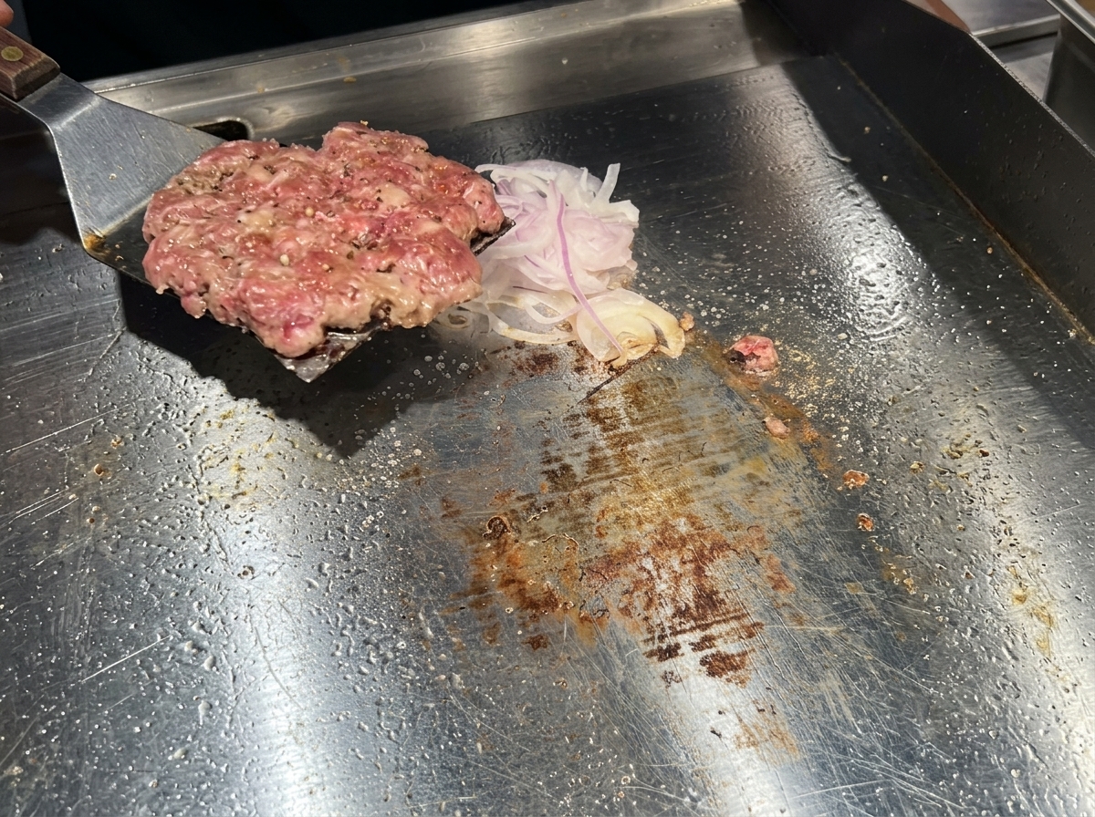 Ein Metallwender wendet das braun gebratene Patty auf die Zwiebelseite um.