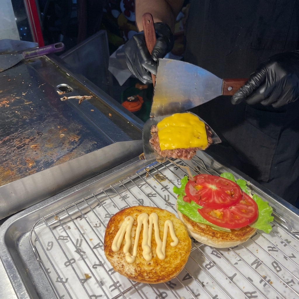 Ein Pfannenwender hebt das Patty mit geschmolzenem Käse auf das vorbereitete Burger-Brötchen.
