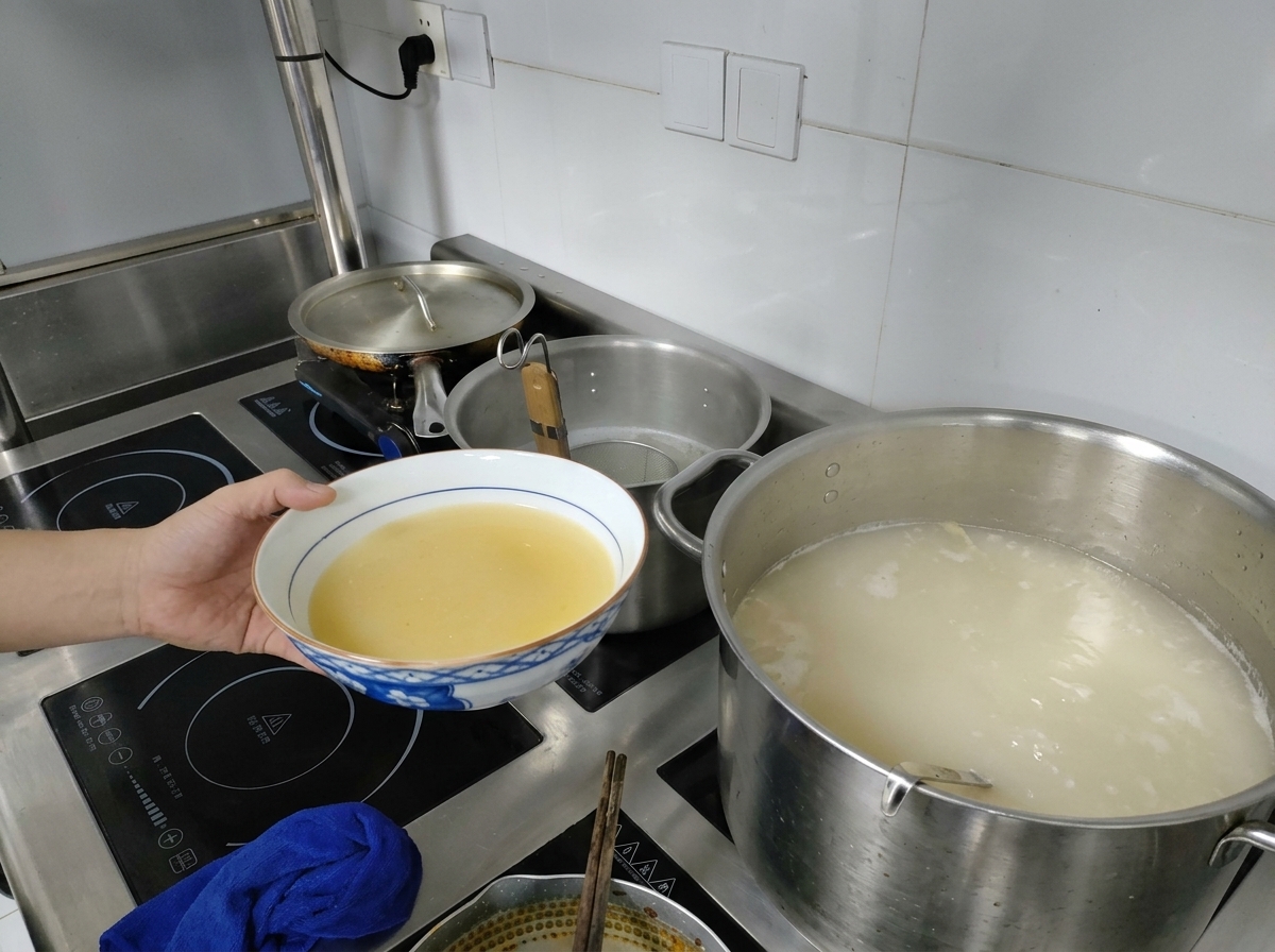 Eine Hand hält eine Schüssel mit gelber Miso-Basis neben einem großen Topf mit kochender, milchig-weißer Brühe auf dem Herd.