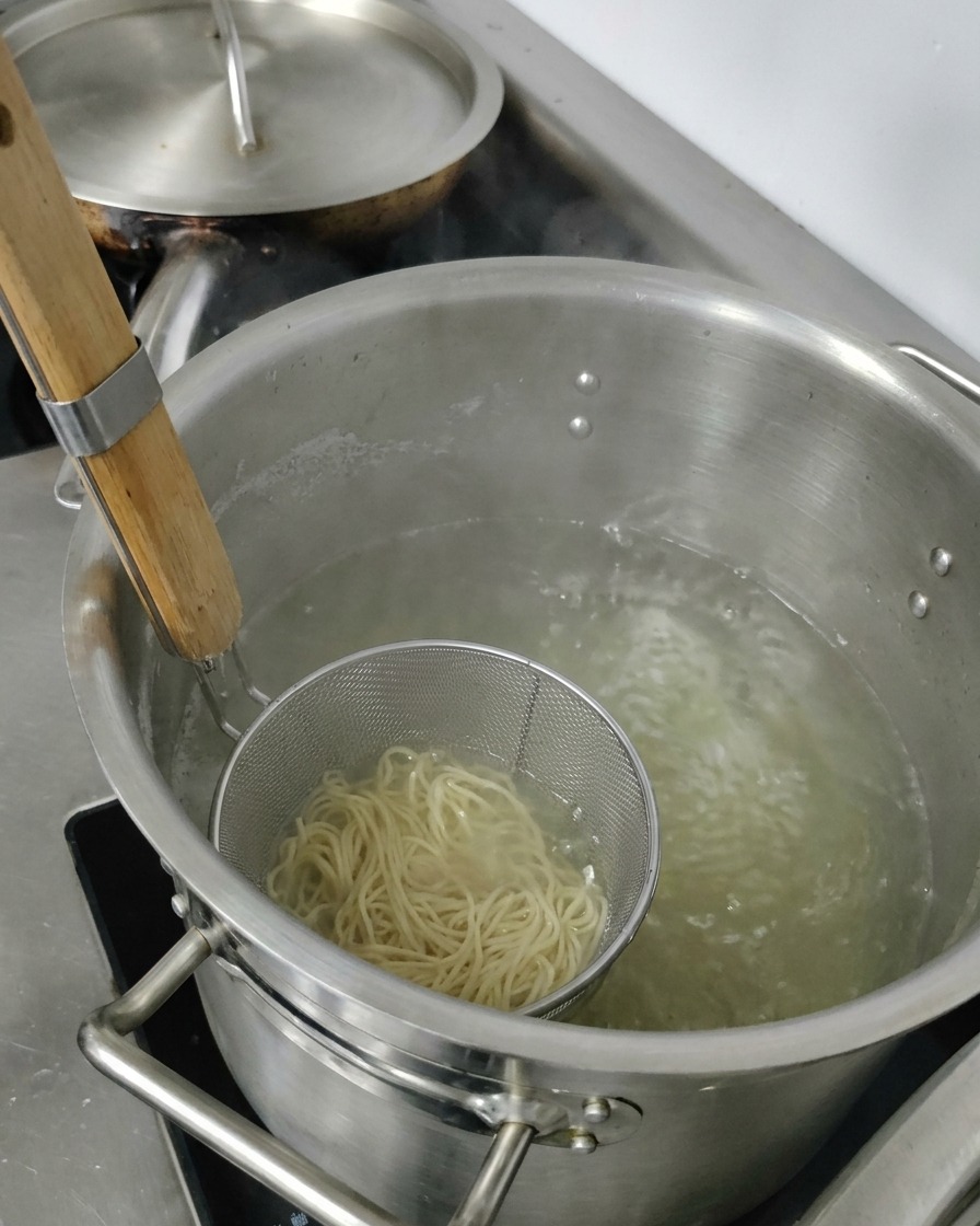 Ein Metallsieb mit gekochten Ramen-Nudeln wird aus einem Topf mit heißem Wasser gehoben.