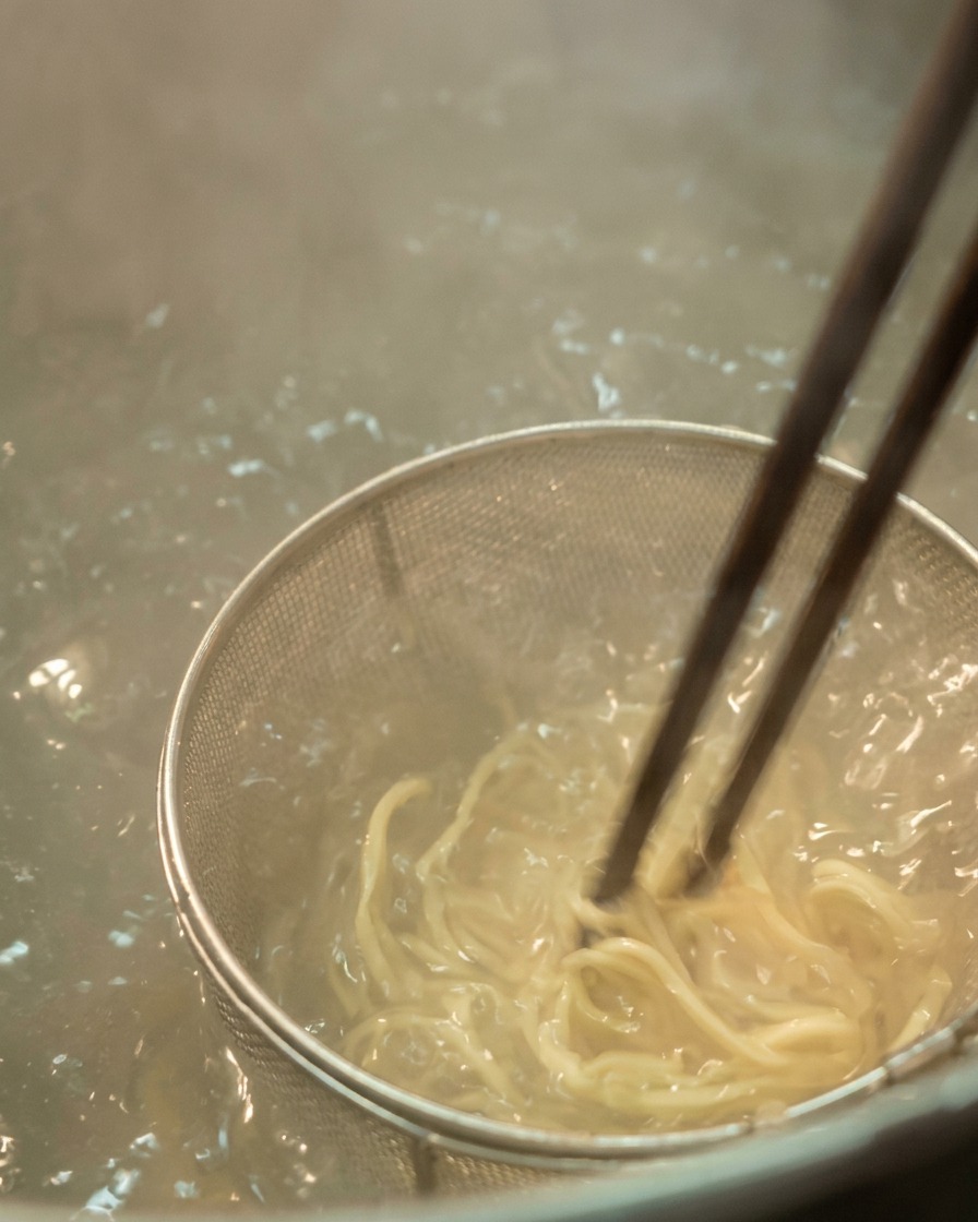 Nahaufnahme von hellen Ramen-Nudeln, die mit dunklen Stäbchen in sprudelnd kochendem Wasser gerührt werden.