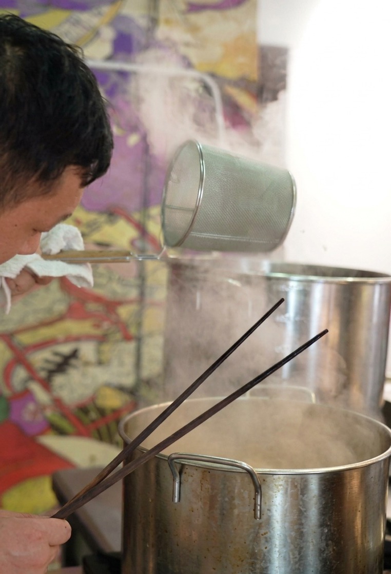Ein Koch schüttelt ein zylindrisches Nudelsieb kräftig über einem Topf, um Wasser aus den Ramen-Nudeln zu entfernen.