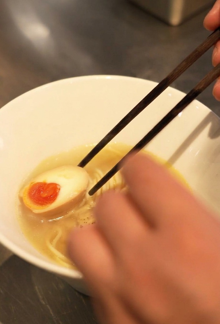 Stäbchen legen ein halbiertes Ei mit flüssigem, orangefarbenem Dotter in die Schüssel mit Ramen.