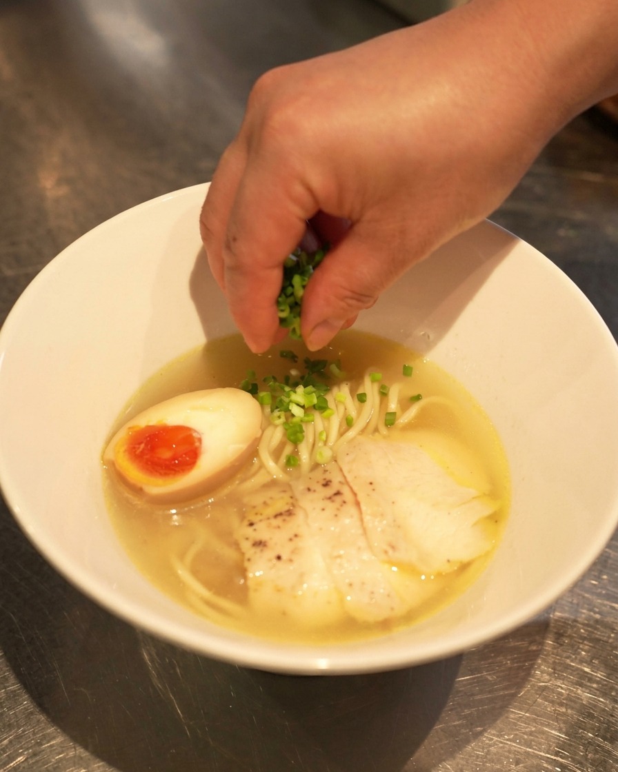 Eine Hand streut frisch gehackte Frühlingszwiebeln über die fertige Schüssel Chicken Shio Ramen.