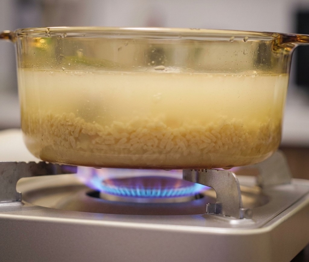 Ein Glastopf mit trüber, gelblicher Ramen-Brühe köchelt über einer blauen Gasflamme auf einem tragbaren Kocher.
