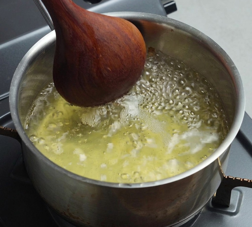 Ein Edelstahltopf mit kochender, heller Brühe wird mit einem dunklen Holzlöffel umgerührt.