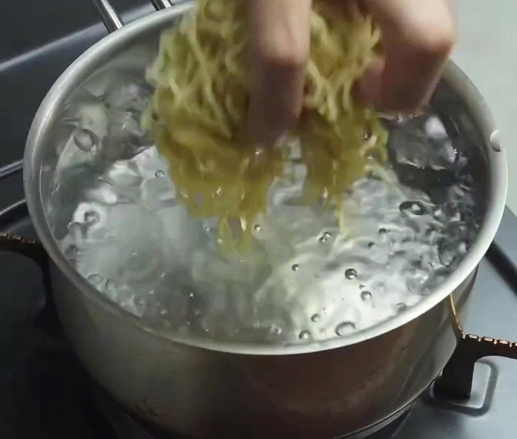 Eine Hand gibt ein Bündel dünner, gewellter Ramen-Nudeln in einen Topf mit sprudelnd kochendem Wasser.