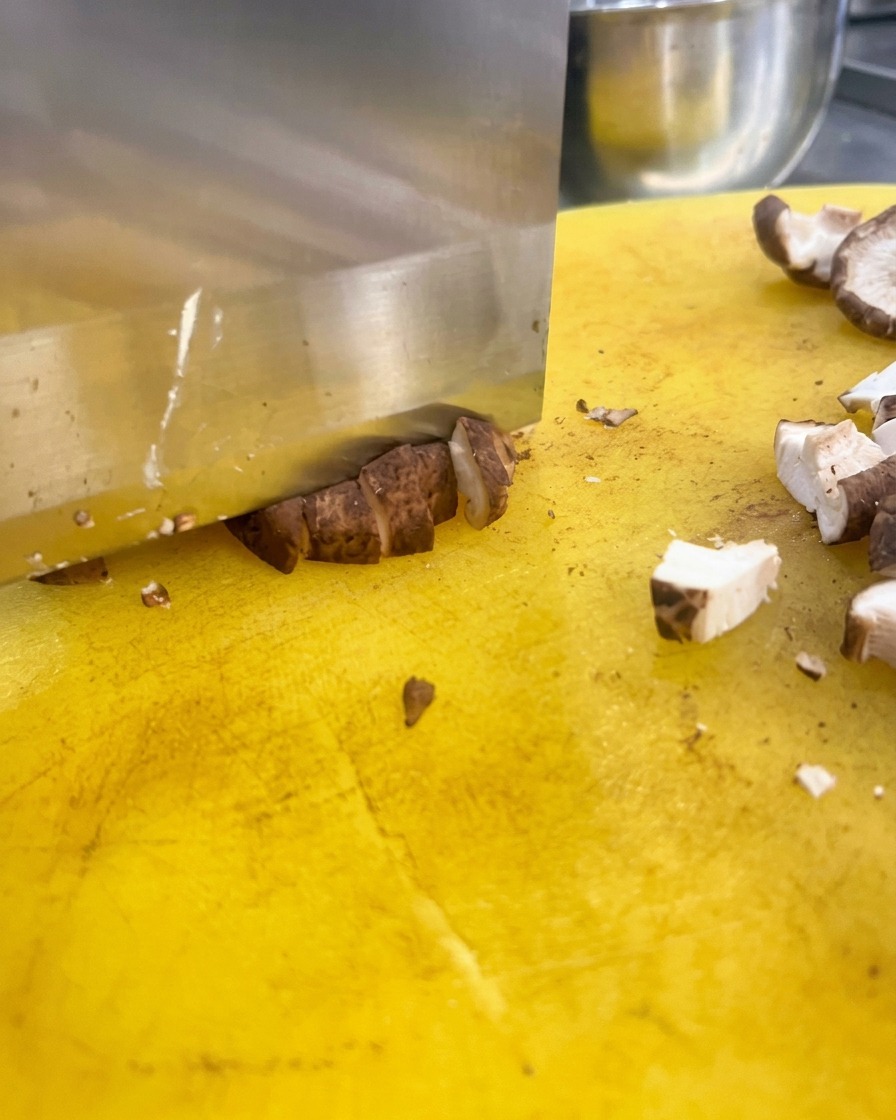 Ein großes Küchenmesser würfelt braune Shiitake-Pilze in kleine Stücke auf einem gelben Schneidebrett.